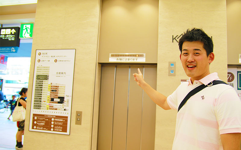 構内のエレベーターを指差すMさん