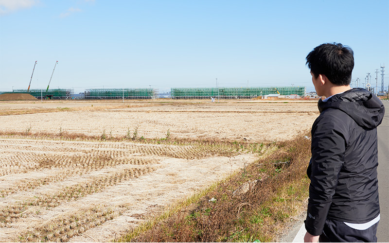 田んぼの向こうに広がる工事現場を見つめる取材班Mさん